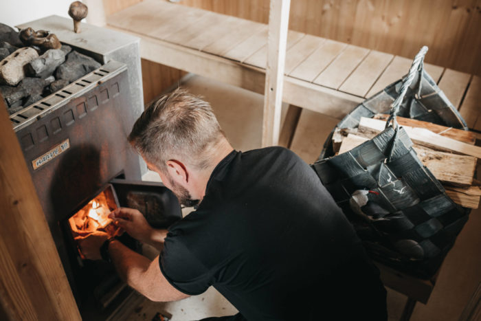 Dans un sauna muni de bancs en bois, un homme est en train de faire du feu dans un poêle en métal de forme rectangulaire surmonté d’un plateau contenant des pierres.