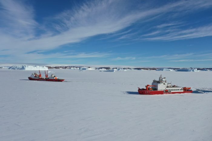 Zwei rote Schiffe fahren in einem schneebedeckten Ozean mit Eisklippen im Hintergrund.