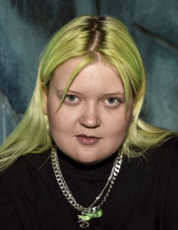 A young woman with her hair dyed green stares into the camera.
