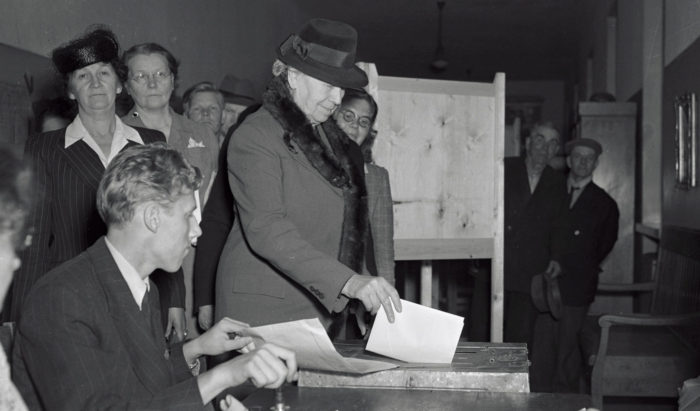 A woman puts a piece of paper through a slot in a box while other people look on.
