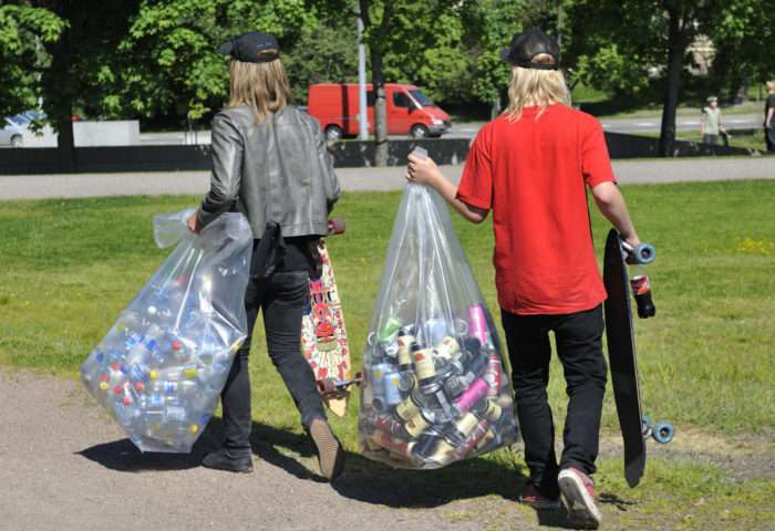Two young men each carry a skateboard in one hand and a large plastic bag filled with bottles and cans in the other hand.