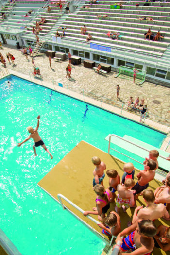 Crianças aguardam sua vez na plataforma de mergulho de 10 metros enquanto uma delas pula na piscina.