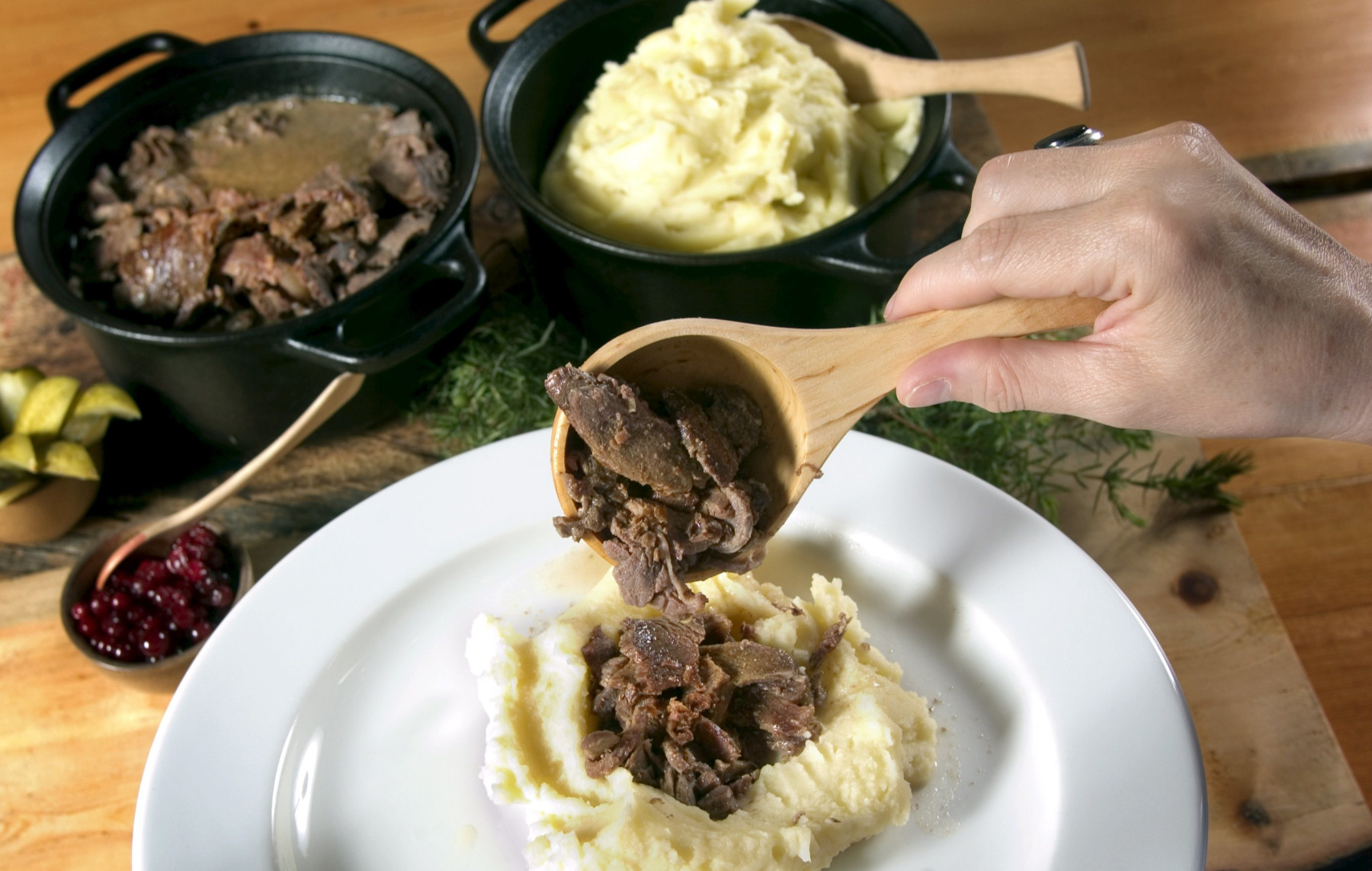 A hand spoons pieces of reindeer meat onto a plate of mashed potatoes.