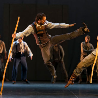 舞台上，一个男人在另一个男人面前跳起并在空中踢腿。