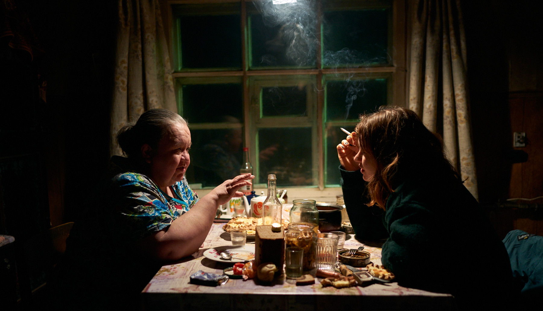 An older woman and a younger woman sit at a kitchen table, drinking and smoking and talking.