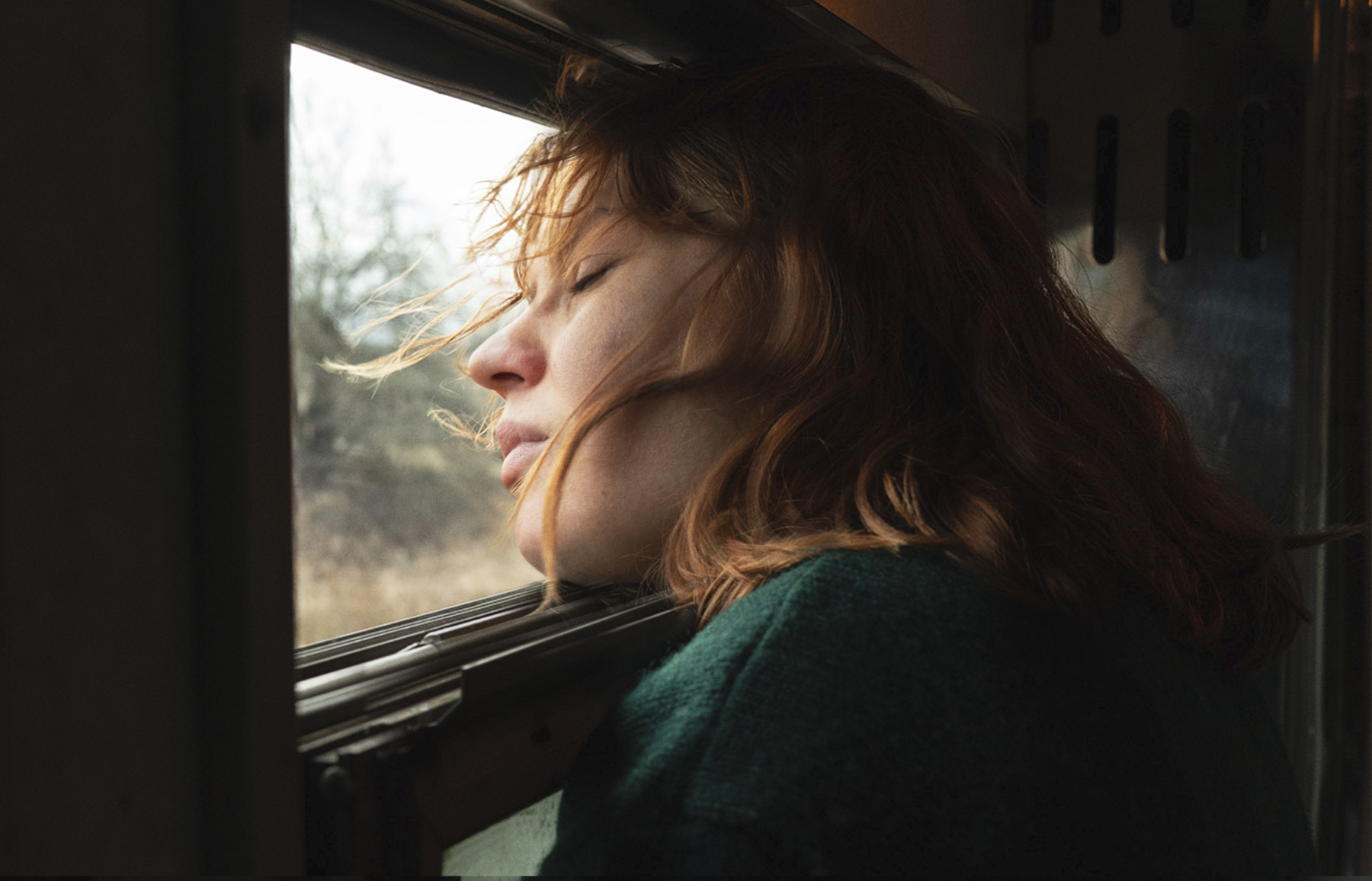 With her eyes closed, a woman lets her hair blow in the breeze at an open train window.