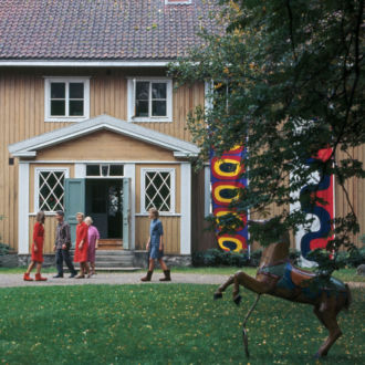 People walk past a country home that has colourful fabric banners hanging from its windows.