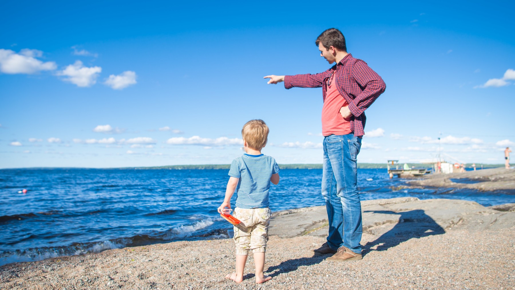 On the shore of a lake, a man points at something on the horizon while a child looks in the same direction.