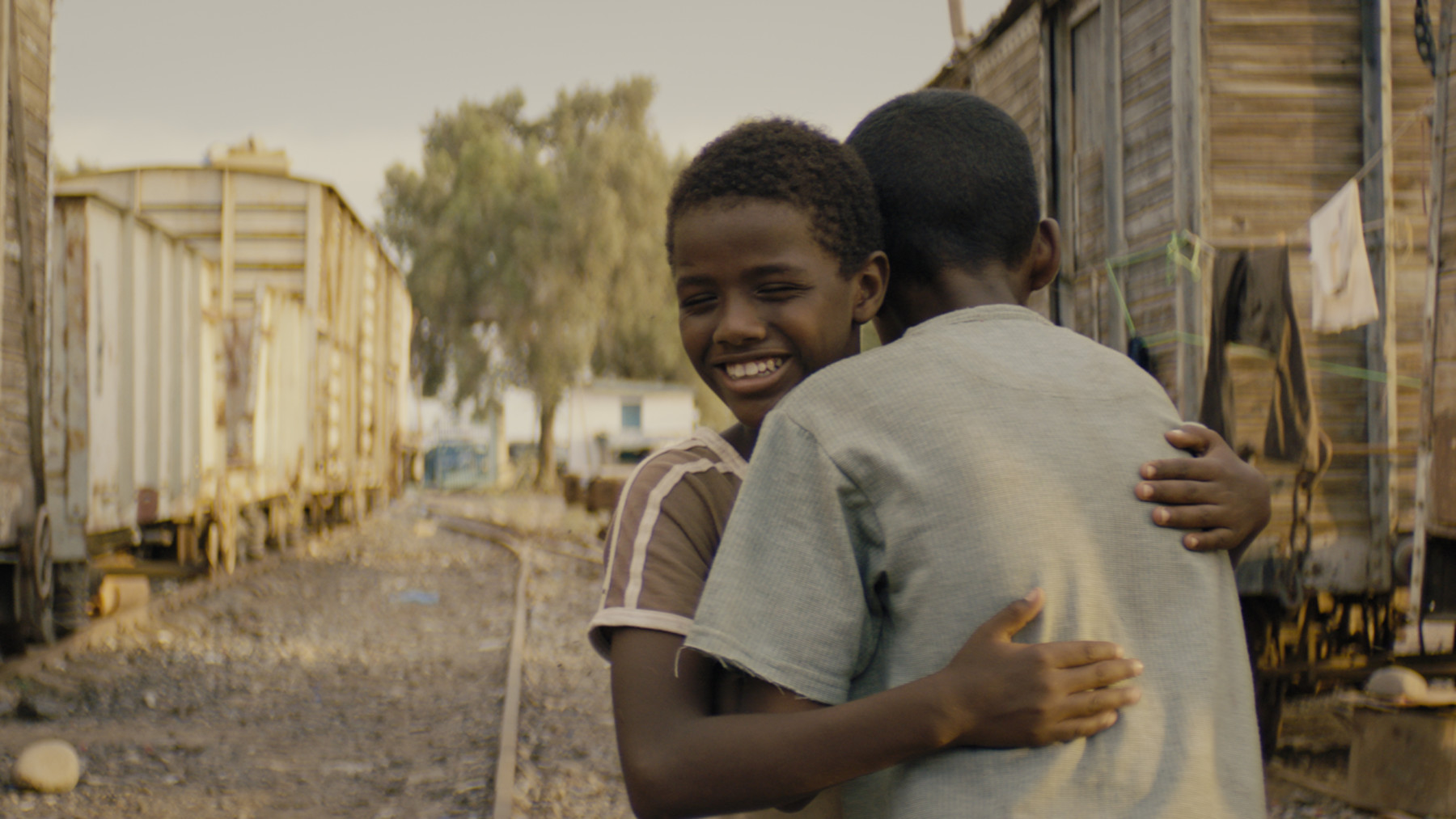 A boy hugs another boy on a gravel street.