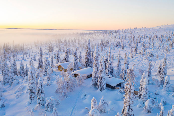 Luftaufnahme einiger Holzhütten in einer verschneiten Waldlandschaft.