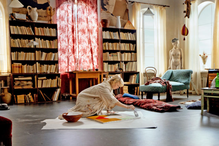 A woman paints on a canvas on the floor of a large room with bookshelves in the background.