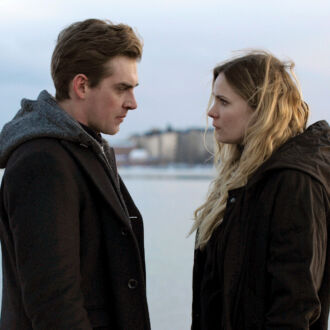 A man and a woman wearing jackets face each other, with a harbor in the background.