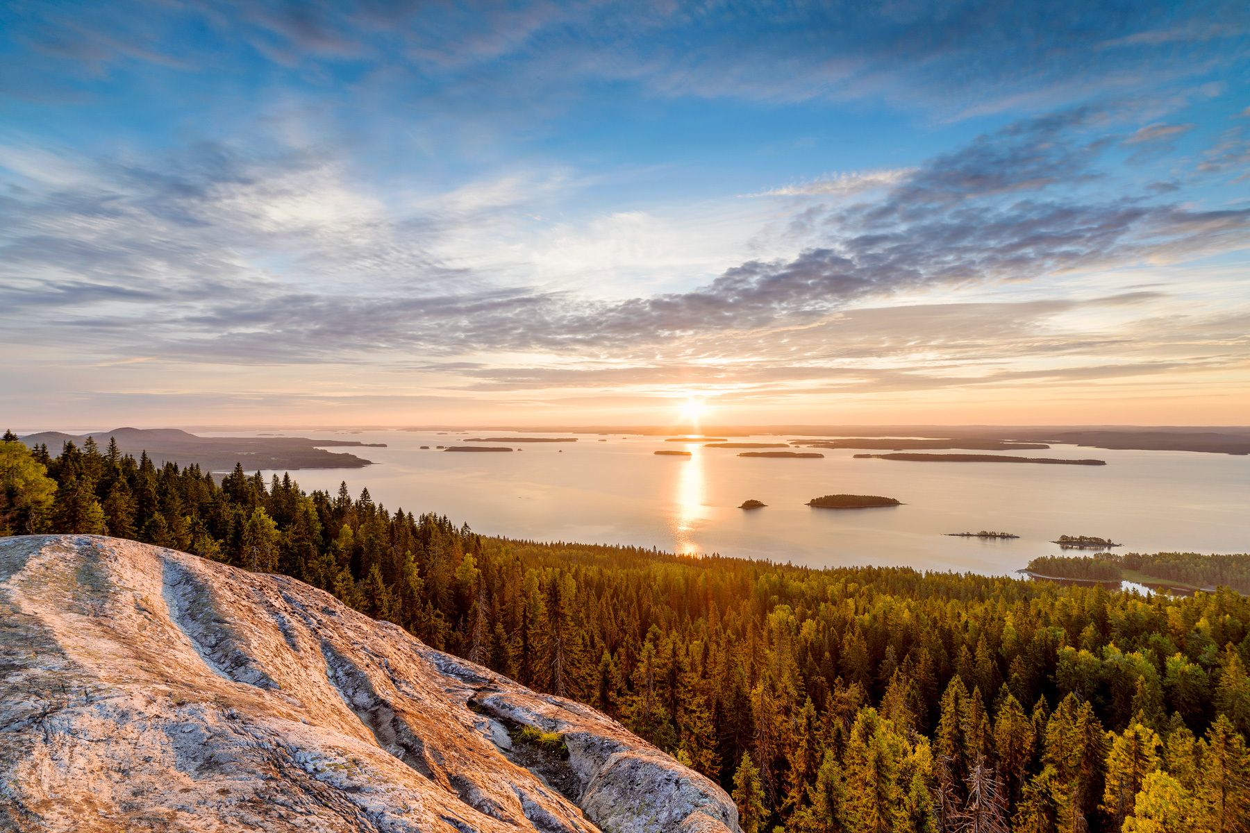 Vue d'un coucher de soleil sur un lac parsemé d'îles depuis une crête surplombant le lac et une forêt.