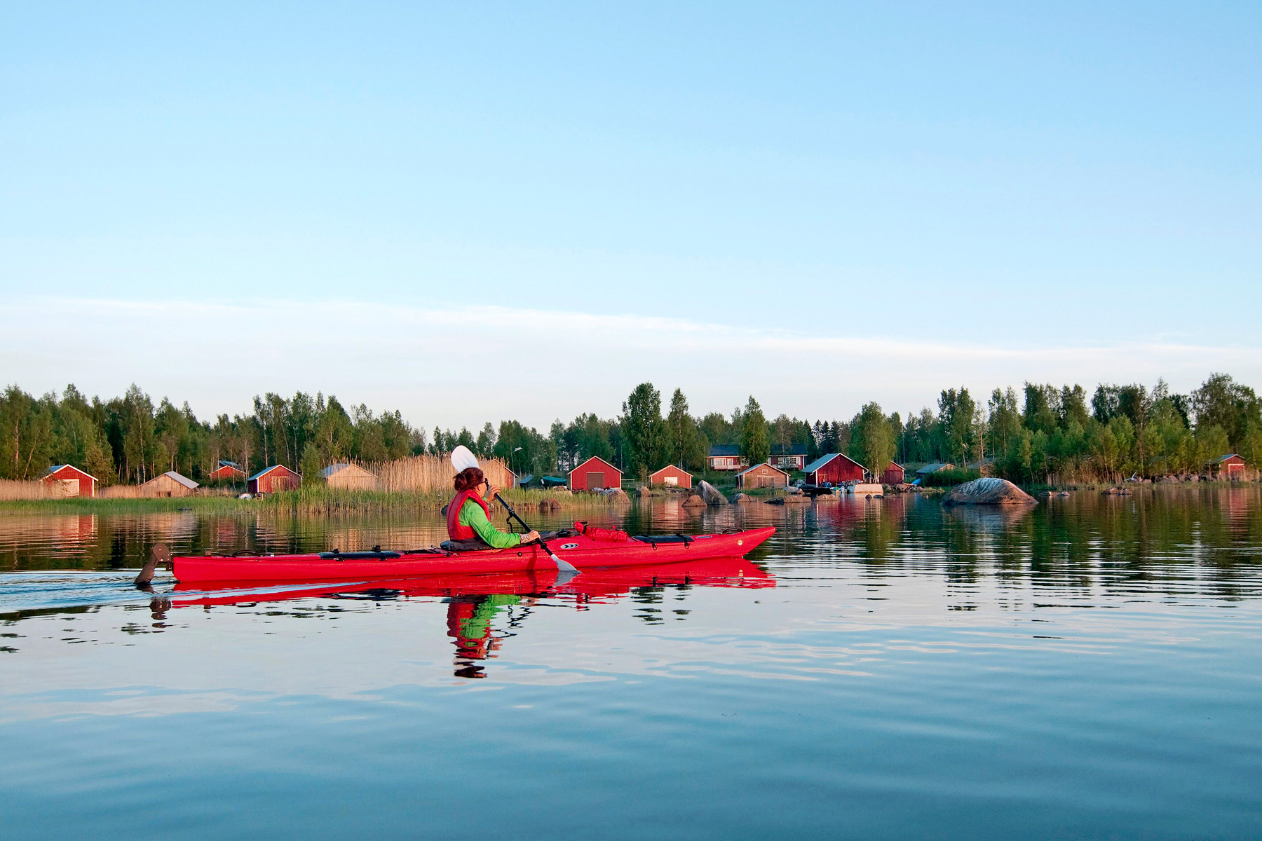 Une personne fait du kayak le long d'un rivage forestier parsemé de hangars à bateaux en bois rouge.