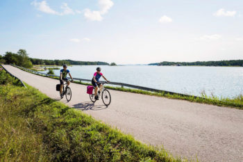 Zwei Personen in sportlicher Kleidung fahren im Sommer mit dem Fahrrad auf einem Küstenweg.