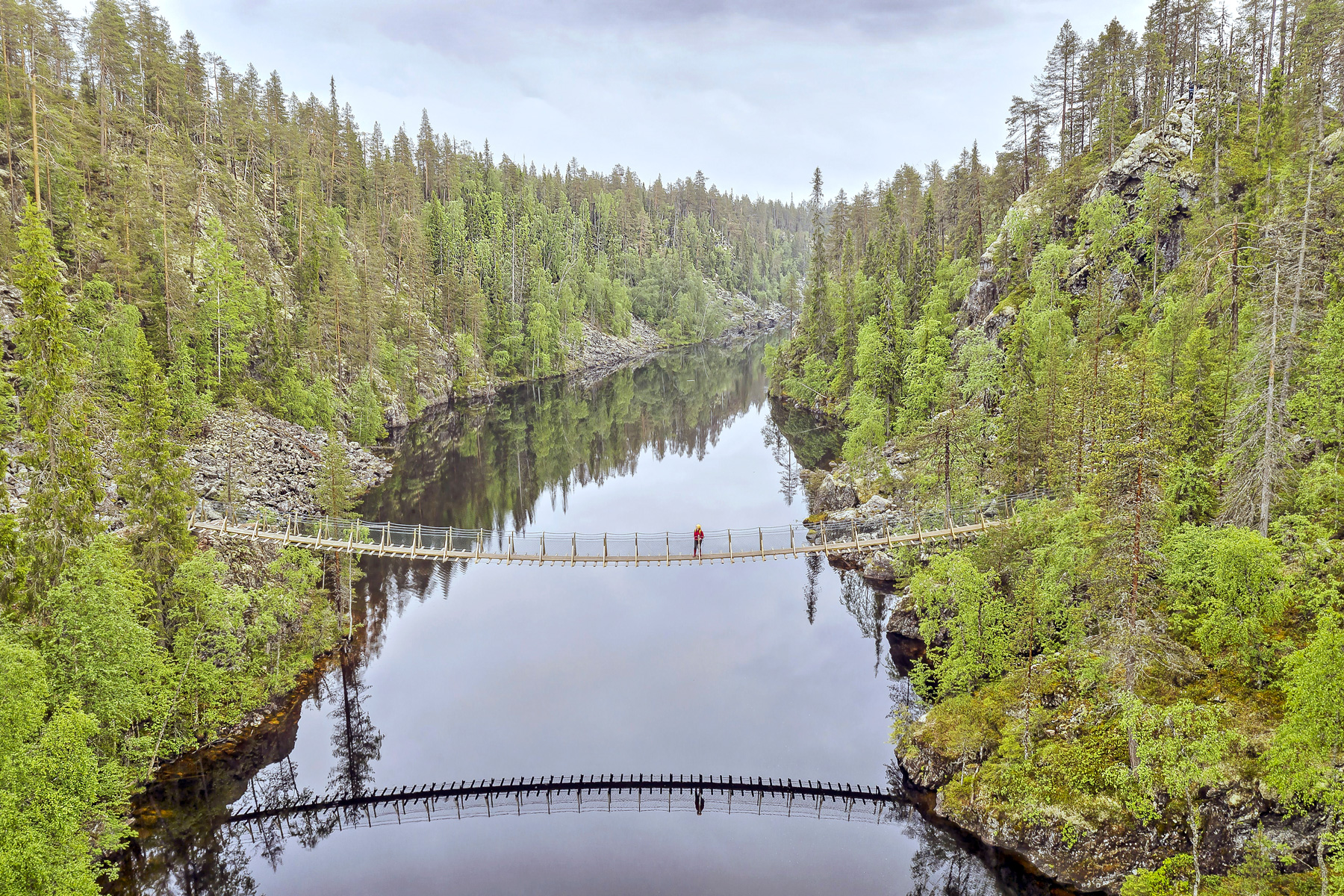 Une personne traverse une large rivière sur un pont suspendu entre deux rives couvertes de forêt.