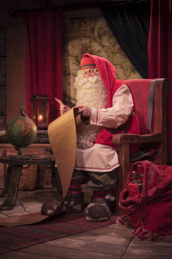 Sitting in a chair, Santa Claus holds a pen in one hand and a long scroll of paper in the other hand.