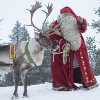 Santa Claus, in a long red coat and a long white beard, stands in a snowy forest with a reindeer.