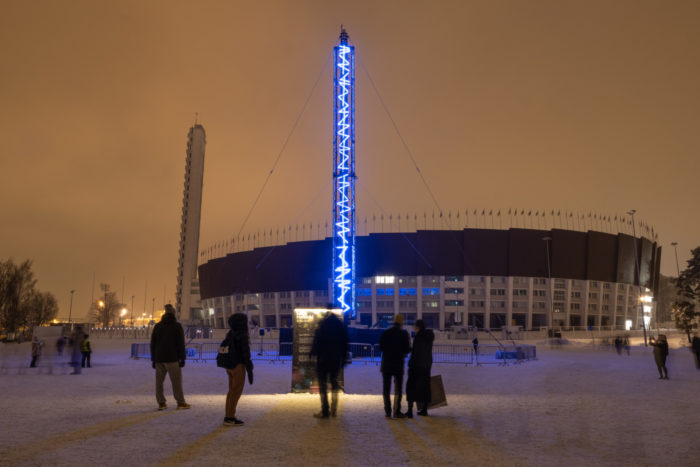 Several people watch a tower with light patterns, in front of a stadium.