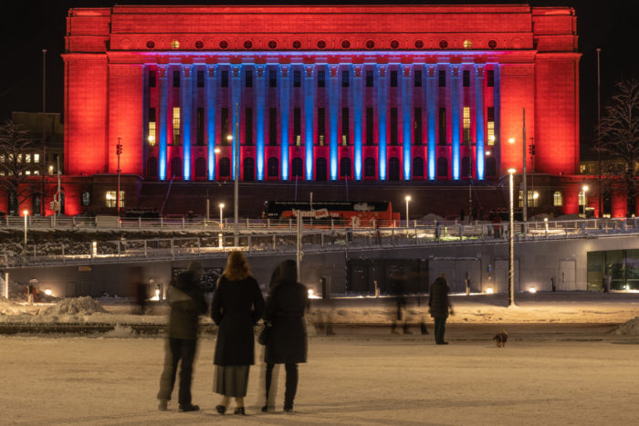 Several people look at the Parliament building, which is lit with various colours of light.