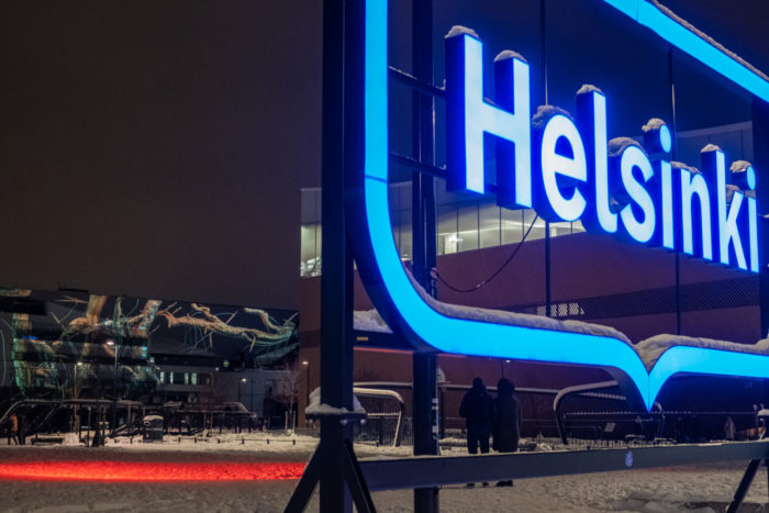 前景中，一座大型灯光标识显示着英文“赫尔辛基”（Helsinki）字样，背景中一件视频投射作品正投射到一座建筑的一侧墙面上。