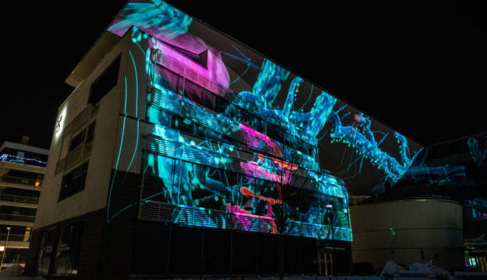 On voit une façade d’immeuble illuminée par la projection vidéo de différentes formes indéfinies.