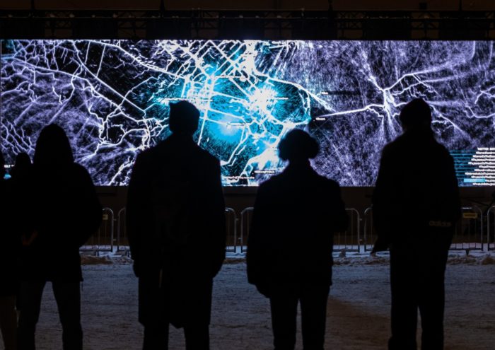 Várias pessoas estão em silhueta na frente de uma tela ampla mostrando uma rede que se assemelha a estradas em um mapa.