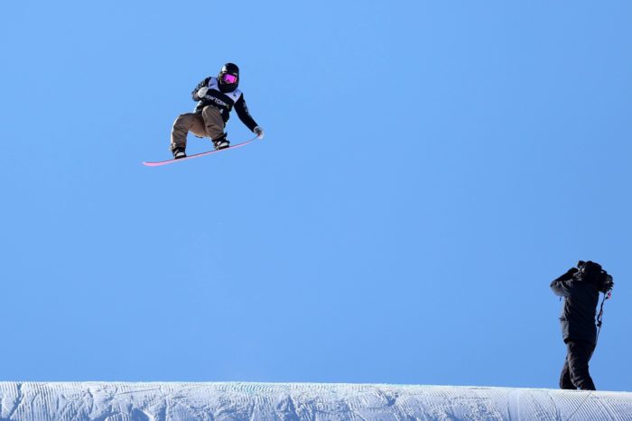 Une snowboardeuse fend les airs, survolant de haut une pente montagneuse enneigée.