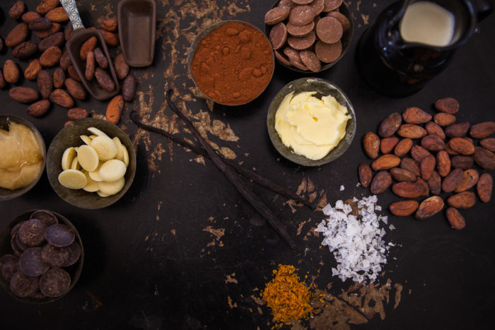 Auf einem Tisch sind eine Reihe von Zutaten für die Schokoladenherstellung angeordnet, darunter Butter, Honig, Sahne, Salz, Vanilleschoten, Kakaopulver und Kakaobohnen.