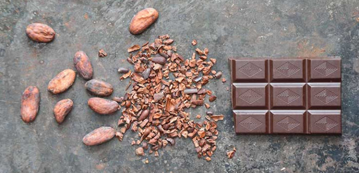 Several whole cocoa beans are arranged next to a pile of crushed cocoa beans and a bar of chocolate.