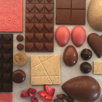 Un assortiment de tablettes de chocolat, d’œufs en chocolat, de fèves de cacao et de baies de toutes les couleurs est présenté sur une table. 