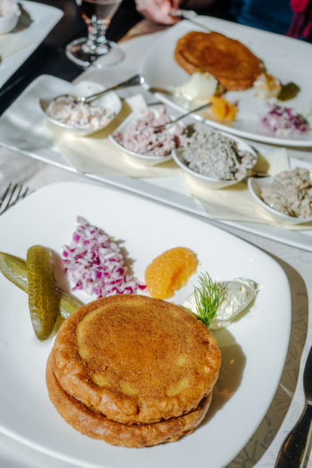 Two plates hold small round pancakes flanked by pickles and other garnishes.