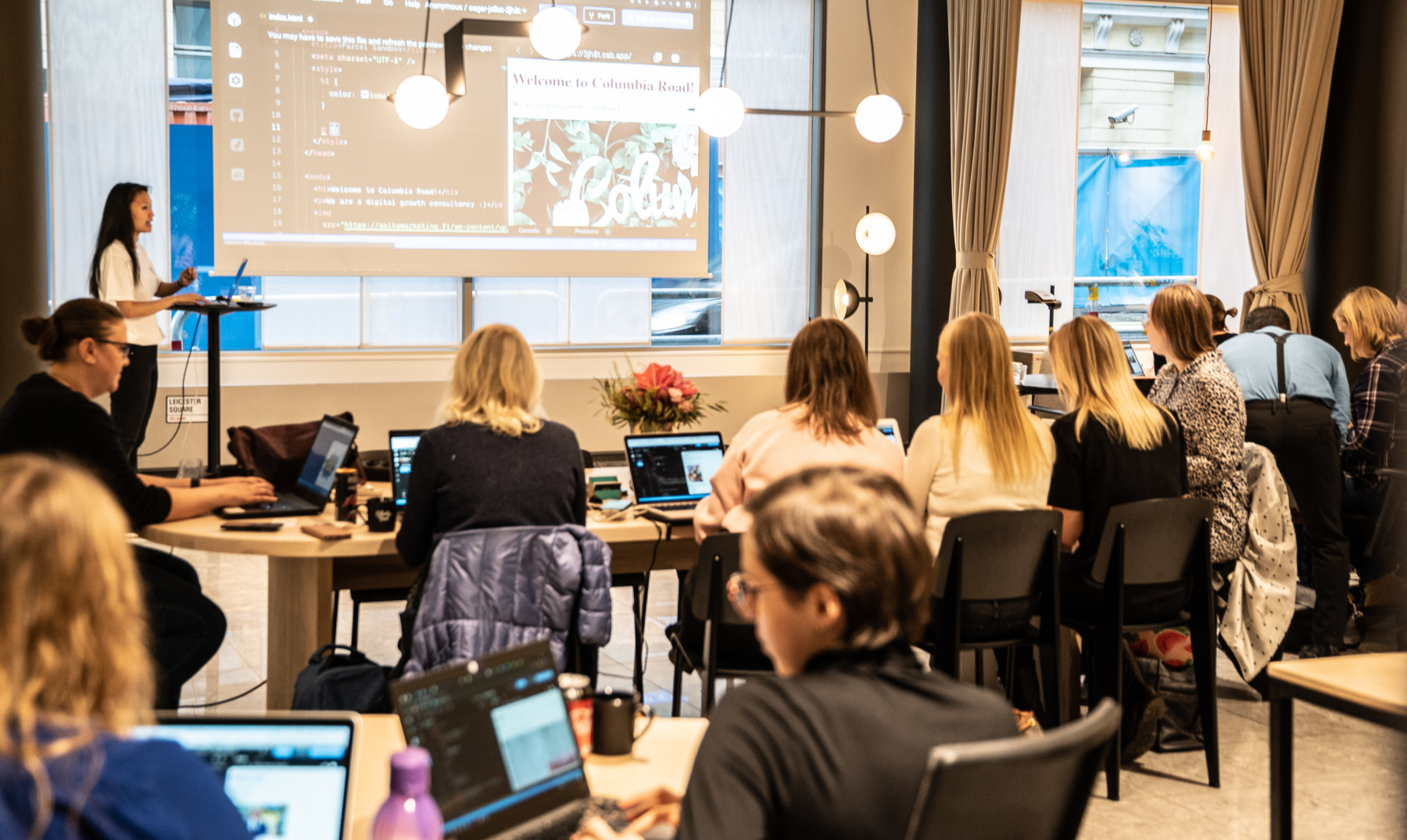 Com seus computadores abertos, um grupo de mulheres observa outra mulher fazer uma apresentação.