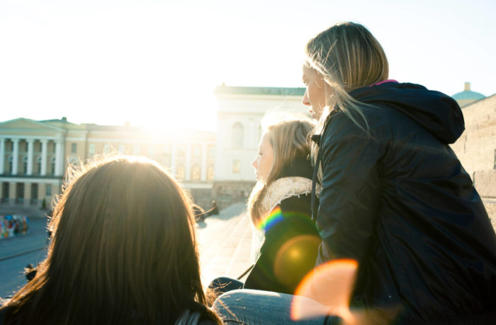 Plusieurs jeunes femmes dont on aperçoit essentiellement la silhouette en raison de l’angle du soleil observent une vaste place urbaine située en contrebas.