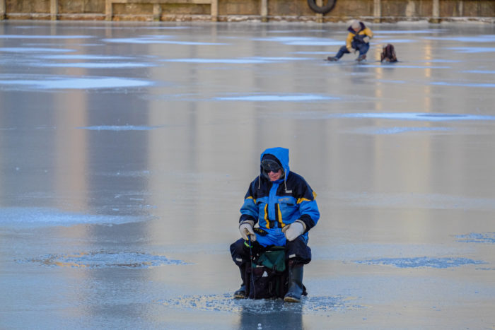 Dos personas con abundante ropa de invierno pescan sentadas en taburetes sobre la superficie del hielo.