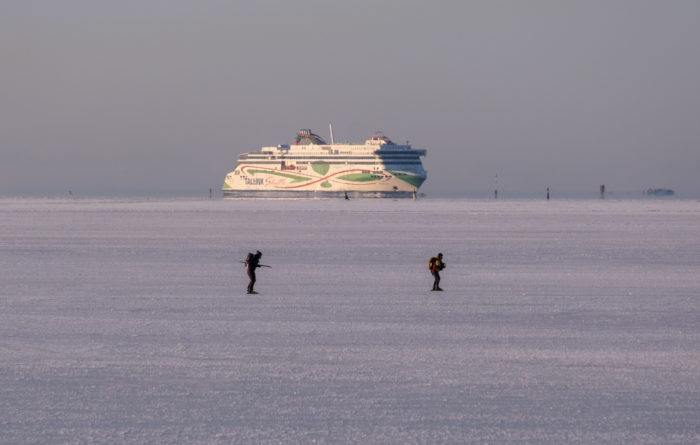 Two small figures are silhouetted in the middle of a wide expanse of sea ice, with a large passenger ship in the background.