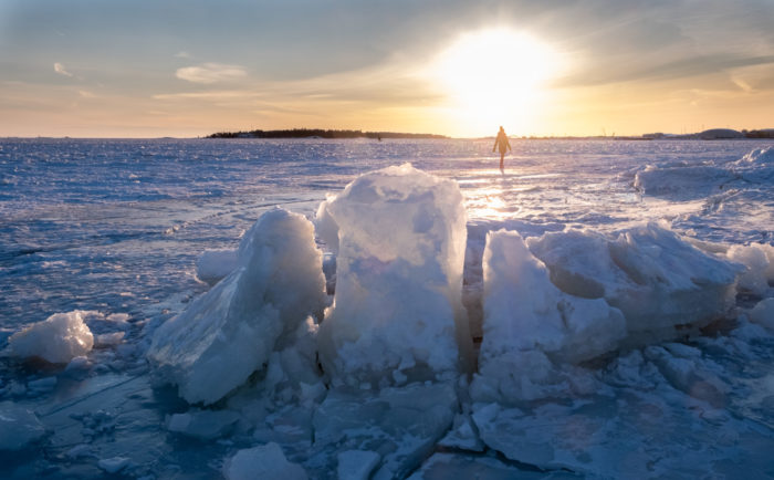 A person walks in front of islands and the setting sun on a wide expanse of sea ice.