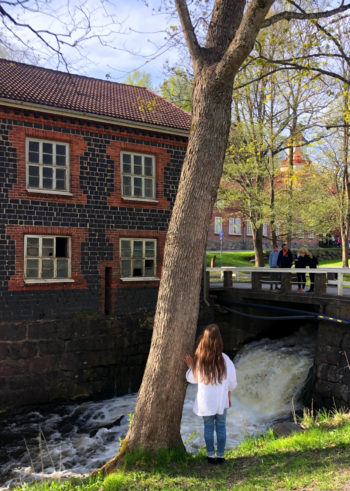 A girl standing by a tree looking at the river.