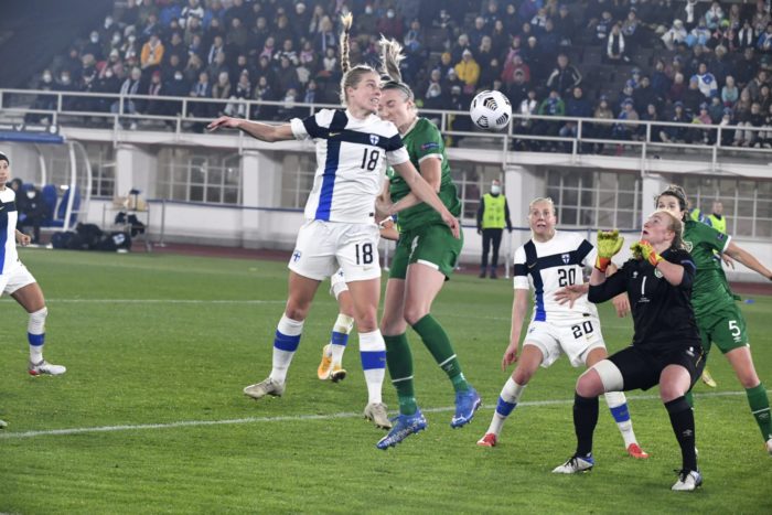 A Finnish soccer player heads the ball as the Irish goalkeeper tries to block it.