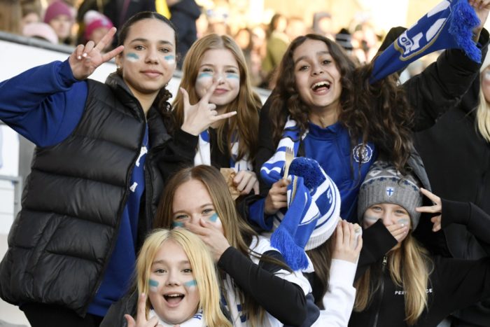 Группа девушек с нарисованными на их щеках синими и белыми полосками аплодирует и машет руками.