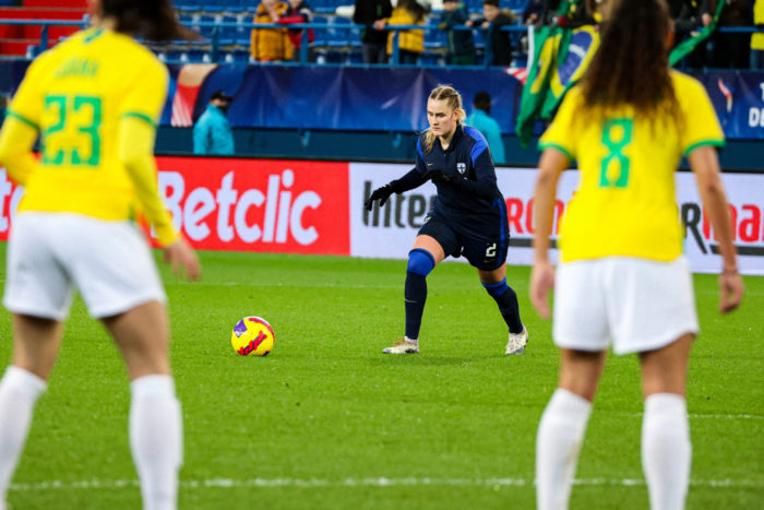 Duas jogadoras de futebol brasileiras ficam de pé enquanto uma finlandesa se prepara para chutar a bola.