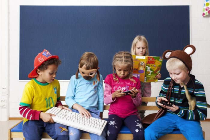 Five kids at a daycare each have a book, a computer keyboard or a mobile device in their hands.