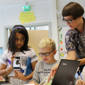 تتحدث معلمة مع أربعة من التلاميذ في تمرين يستخدمون فيه كتابًا، وجهاز كمبيوتر محمولاً، وهاتفًا محمولاً.