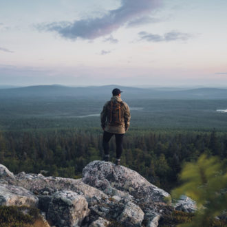 A man stands on a rock, looking out over a view of forests and lakes.