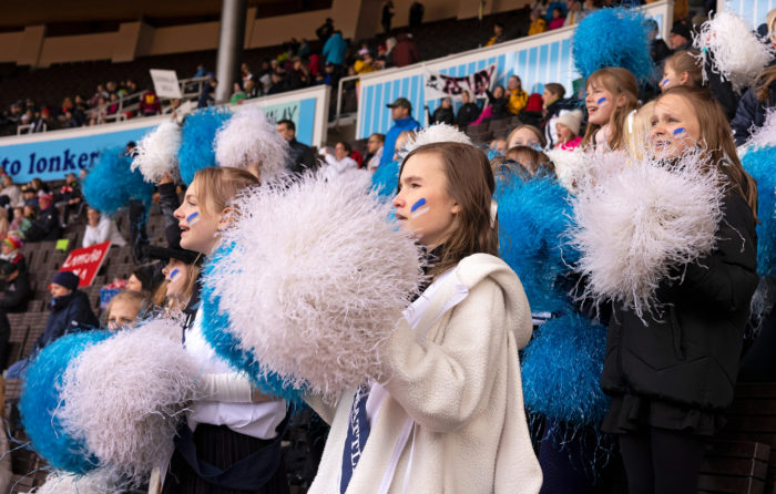 A group of children wave blue and white pom-poms and cheer.