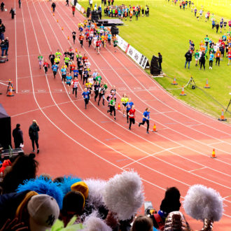 A group of children runs on a track in a stadium with an audience watching.