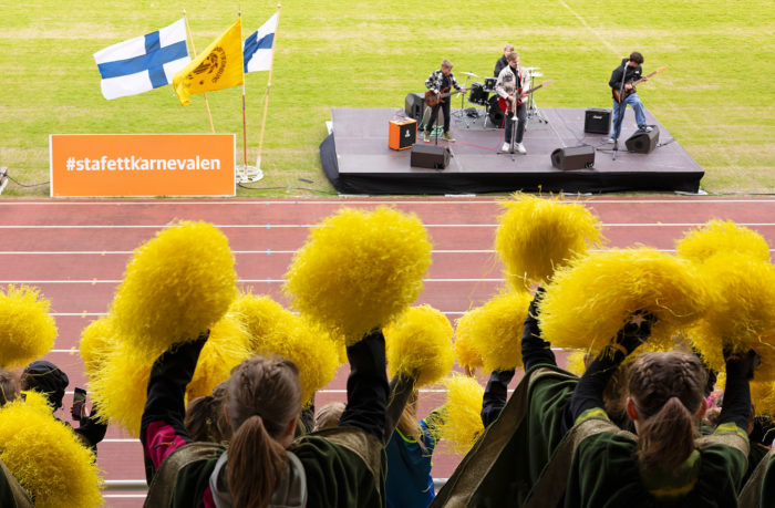 A band plays on a stage beside a running track, and cheerleaders wave yellow pom-poms.