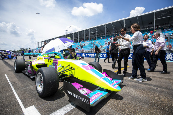 À côté d’une voiture à l’arrêt sur un circuit automobile, une personne tient une ombrelle au-dessus de la pilote pour la protéger du soleil tandis que des gens discutent entre eux tout en désignant le véhicule du doigt.