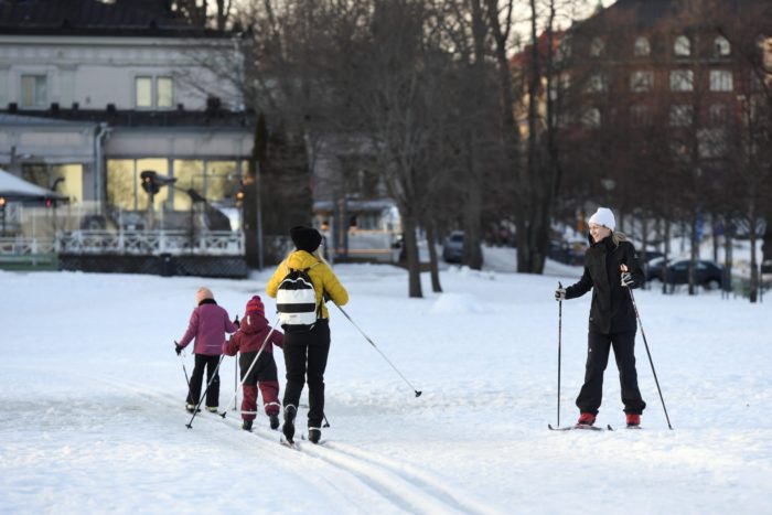 Dois adultos e duas crianças praticam esqui cross-country em um parque com prédios da cidade visíveis ao fundo.