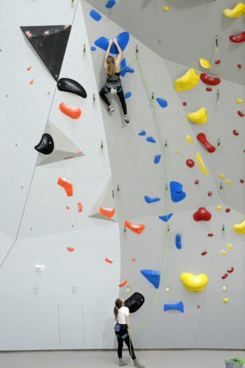 A woman climbs high up on a climbing wall while another woman holds the safety rope for her.